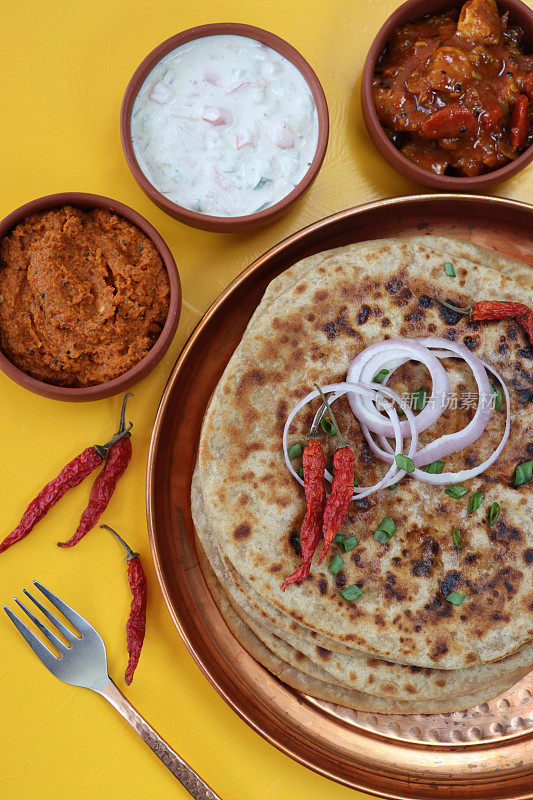 金属托盘上放着aloo paratha，上面撒着辣椒、红洋葱圈和切碎的韭菜，美味的面饼里塞满了捣碎的garam masala土豆、酸辣酱、泡菜蘸酱和raita(酸奶)，俯瞰美景